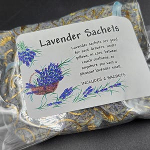 Puede incluir: Una bolsa de pl&aacute;stico transparente que contiene dos saquitos de lavanda. Los saquitos est&aacute;n decorados con un estampado floral dorado y azul. Una etiqueta blanca muestra el texto "Saquitos de lavanda" y una descripci&oacute;n del producto, con un dibujo de lavanda.