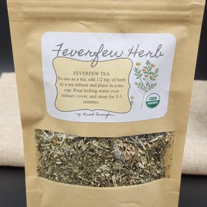 Puede incluir: Una bolsa de papel marr&oacute;n con una ventana transparente que muestra hierba de matricaria seca. La etiqueta dice "Feverfew Herb" e incluye instrucciones para hacer t&eacute;. La bolsa es USDA Organic.