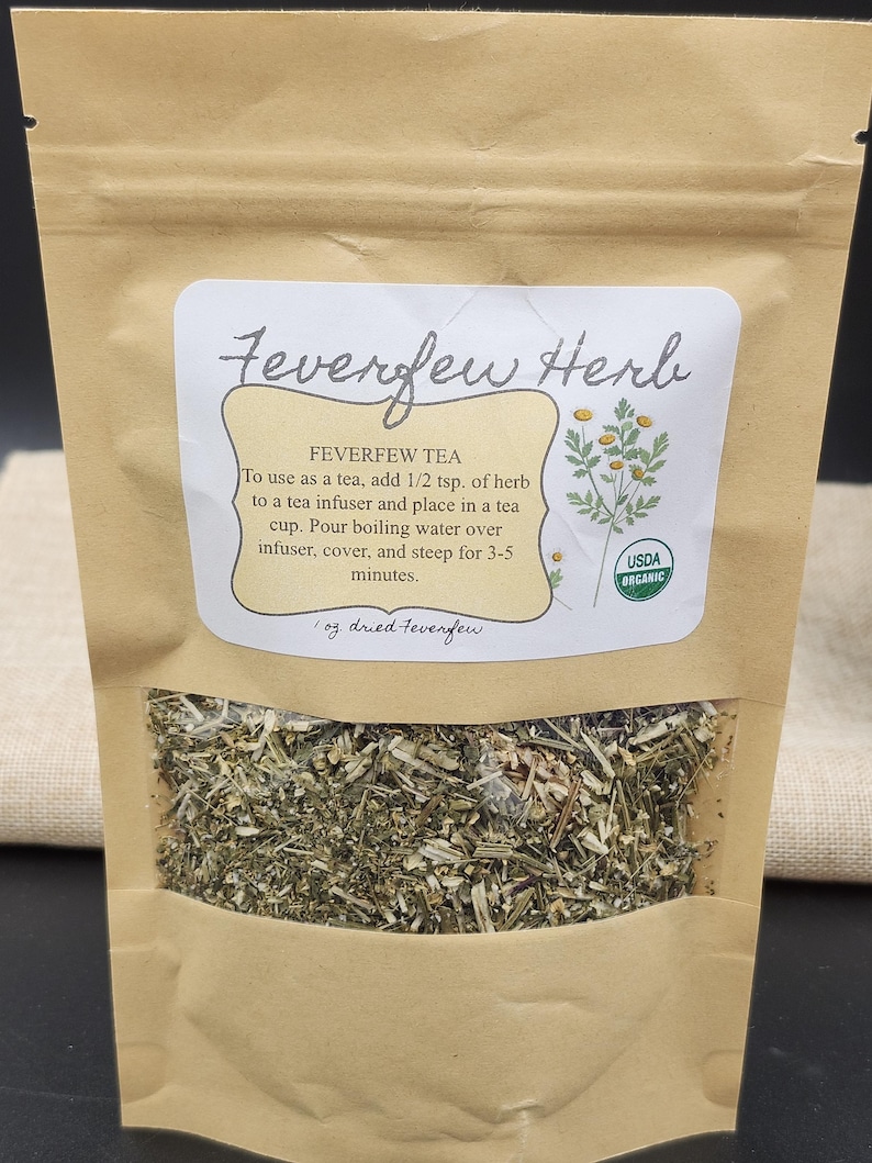 Puede incluir: Una bolsa de papel marr&oacute;n con una ventana transparente que muestra hierba de matricaria seca. La etiqueta dice "Feverfew Herb" e incluye instrucciones para hacer t&eacute;. La bolsa es USDA Organic.