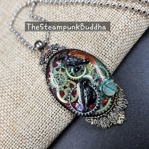 Peut inclure: Collier pendentif de style steampunk avec une cha&icirc;ne argent&eacute;e. Le pendentif ovale pr&eacute;sente un design d&eacute;taill&eacute; avec des engrenages, un corbeau et le texte "TheSteampunkBuddha". Le pendentif a un code couleur rouge, or et sarcelle.
