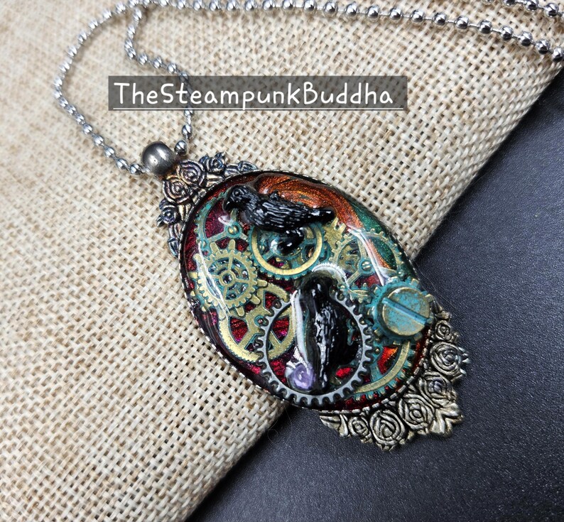 Peut inclure: Collier pendentif de style steampunk avec une cha&icirc;ne argent&eacute;e. Le pendentif ovale pr&eacute;sente un design d&eacute;taill&eacute; avec des engrenages, un corbeau et le texte "TheSteampunkBuddha". Le pendentif a un code couleur rouge, or et sarcelle.