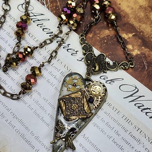 May include: Steampunk-style heart pendant necklace. The pendant features gears, filigree, and a dragonfly, with a mix of gold, bronze, and purple accents. The necklace has a chain with faceted beads.