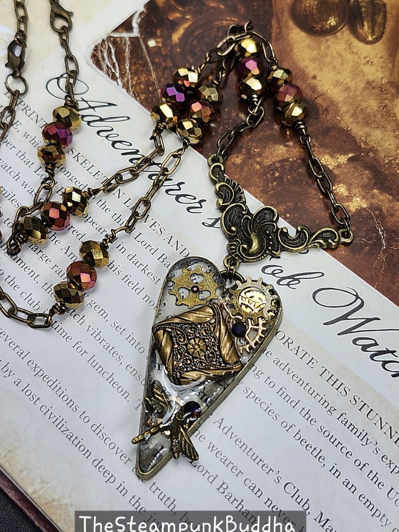May include: Steampunk-style heart pendant necklace. The pendant features gears, filigree, and a dragonfly, with a mix of gold, bronze, and purple accents. The necklace has a chain with faceted beads.