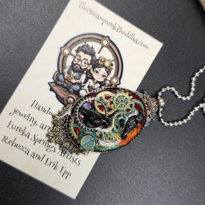 Peut inclure: Un collier pendentif ovale avec un design steampunk. Le pendentif pr&eacute;sente des engrenages, un oiseau noir et des accents color&eacute;s. Le collier a une cha&icirc;ne argent&eacute;e et une bordure d&eacute;corative. La carte de visite indique "TheSteampunkBuddha.com".