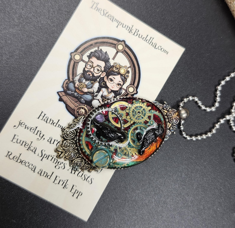 Peut inclure: Un collier pendentif ovale avec un design steampunk. Le pendentif pr&eacute;sente des engrenages, un oiseau noir et des accents color&eacute;s. Le collier a une cha&icirc;ne argent&eacute;e et une bordure d&eacute;corative. La carte de visite indique "TheSteampunkBuddha.com".