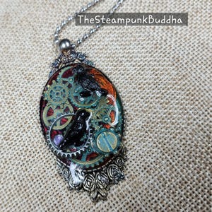 Peut inclure: Collier pendentif ovale avec un design steampunk. Le pendentif pr&eacute;sente deux oiseaux noirs, des engrenages et une gemme violette sur un fond rouge, orange et turquoise. Le pendentif est encadr&eacute; d'un m&eacute;tal argent&eacute; et est suspendu &agrave; une cha&icirc;ne argent&eacute;e.