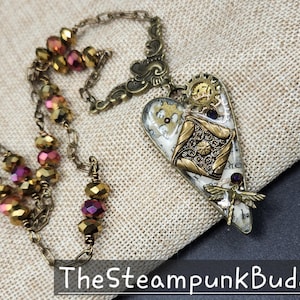 May include: A steampunk-style necklace with a heart-shaped pendant. The pendant features gears, a dragonfly, and a decorative design in gold and silver tones. The necklace has a chain with faceted beads in shades of gold and purple.