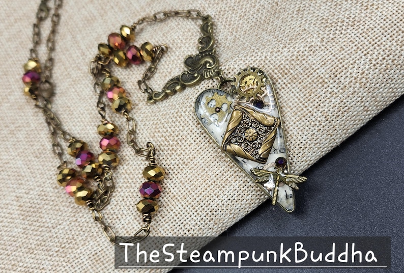 May include: A steampunk-style necklace with a heart-shaped pendant. The pendant features gears, a dragonfly, and a decorative design in gold and silver tones. The necklace has a chain with faceted beads in shades of gold and purple.