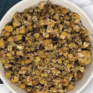 K&ouml;nnte beinhalten: Eine wei&szlig;e Sch&uuml;ssel gef&uuml;llt mit getrockneten Kamillenbl&uuml;ten. Die Bl&uuml;ten sind eine Mischung aus Gelb und Braun, mit kleinen, zarten Bl&uuml;tenbl&auml;ttern und St&auml;ngeln. Das Bild ist eine Nahaufnahme, die die Textur und die Details der getrockneten Kr&auml;uter hervorhebt.