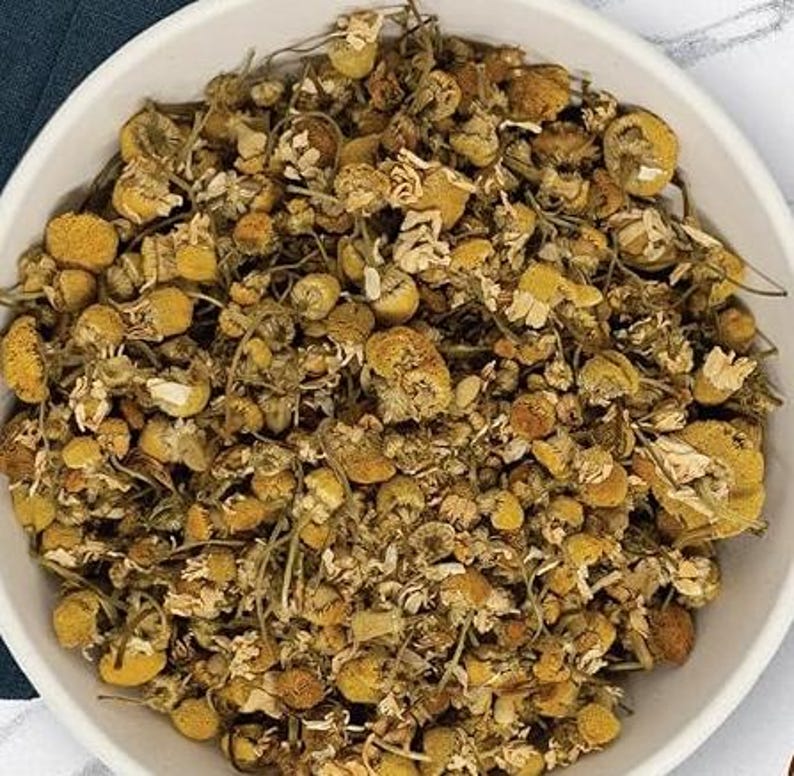 K&ouml;nnte beinhalten: Eine wei&szlig;e Sch&uuml;ssel gef&uuml;llt mit getrockneten Kamillenbl&uuml;ten. Die Bl&uuml;ten sind eine Mischung aus Gelb und Braun, mit kleinen, zarten Bl&uuml;tenbl&auml;ttern und St&auml;ngeln. Das Bild ist eine Nahaufnahme, die die Textur und die Details der getrockneten Kr&auml;uter hervorhebt.