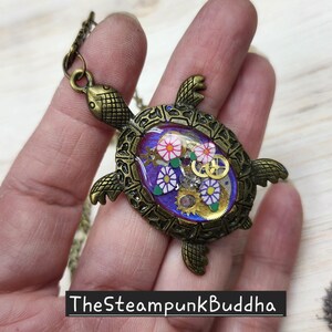 K&ouml;nnte beinhalten: Ein bronzefarbener Schildkr&ouml;ten-Anh&auml;nger mit einer ovalen, violetten Einlage. Der Panzer enth&auml;lt kleine goldfarbene Uhrwerk-Zahnr&auml;der sowie wei&szlig;e und violette blumenf&ouml;rmige Akzente. Der Text "TheSteampunkBuddha" ist in Wei&szlig; auf einem schwarzen Balken gedruckt.