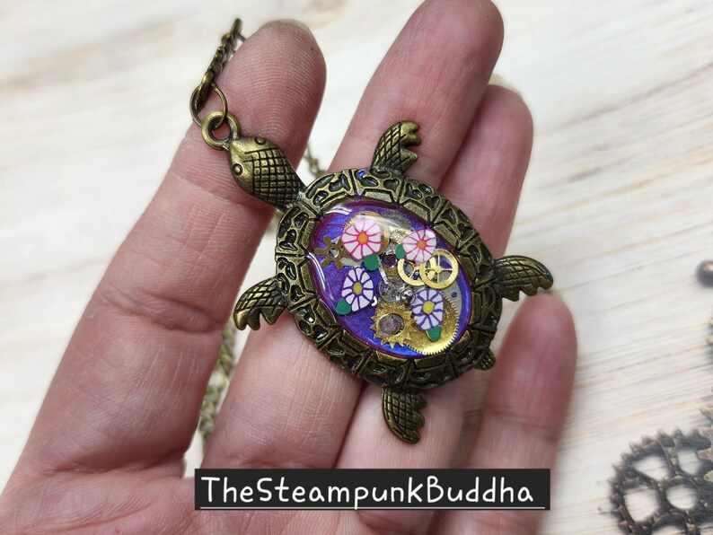 K&ouml;nnte beinhalten: Ein bronzefarbener Schildkr&ouml;ten-Anh&auml;nger mit einer ovalen, violetten Einlage. Der Panzer enth&auml;lt kleine goldfarbene Uhrwerk-Zahnr&auml;der sowie wei&szlig;e und violette blumenf&ouml;rmige Akzente. Der Text "TheSteampunkBuddha" ist in Wei&szlig; auf einem schwarzen Balken gedruckt.