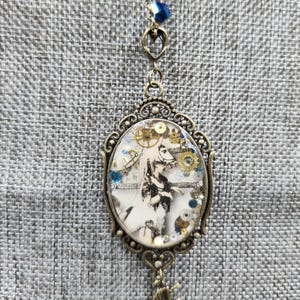 May include: A steampunk-themed pendant necklace. The oval pendant features a black and white image with gears and other small embellishments. The pendant is framed in an ornate, bronze-colored metal. A small unicorn charm hangs below the pendant, and a blue bead accents the chain.