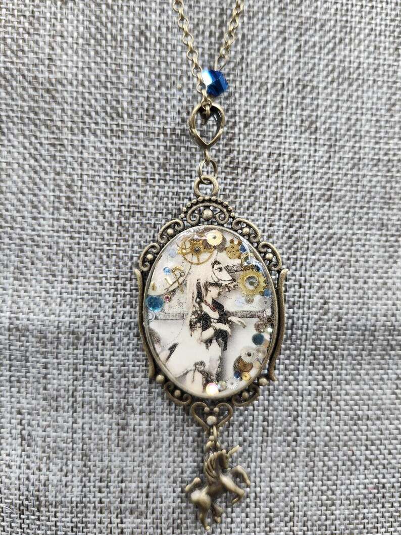 May include: A steampunk-themed pendant necklace. The oval pendant features a black and white image with gears and other small embellishments. The pendant is framed in an ornate, bronze-colored metal. A small unicorn charm hangs below the pendant, and a blue bead accents the chain.