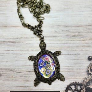 K&ouml;nnte beinhalten: Eine schildkr&ouml;tenf&ouml;rmige Anh&auml;ngerkette in Bronzefarbe mit einer zentralen ovalen Einlage auf blauem Grund, kleinen Blumenmustern und metallischen Zahnradakzenten. Der Panzer weist detaillierte Texturen auf und der Text TheSteampunkBuddha ist sichtbar.