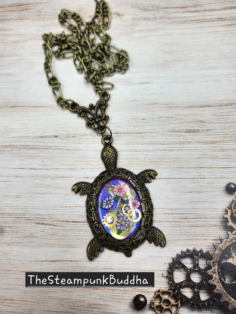 K&ouml;nnte beinhalten: Eine schildkr&ouml;tenf&ouml;rmige Anh&auml;ngerkette in Bronzefarbe mit einer zentralen ovalen Einlage auf blauem Grund, kleinen Blumenmustern und metallischen Zahnradakzenten. Der Panzer weist detaillierte Texturen auf und der Text TheSteampunkBuddha ist sichtbar.