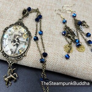 May include: A steampunk-themed jewelry set featuring a pendant with a vintage image and a unicorn charm, along with matching earrings. The set includes blue crystal accents, brass-toned chains, and gear-shaped details. The text "TheSteampunkBuddha" is visible.
