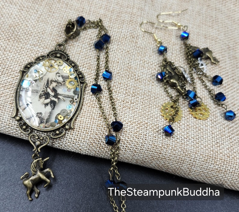 May include: A steampunk-themed jewelry set featuring a pendant with a vintage image and a unicorn charm, along with matching earrings. The set includes blue crystal accents, brass-toned chains, and gear-shaped details. The text "TheSteampunkBuddha" is visible.
