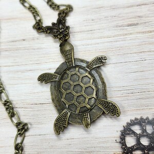 K&ouml;nnte beinhalten: Eine bronzefarbene Metallhalskette mit einem schildkr&ouml;tenf&ouml;rmigen Anh&auml;nger, der ein strukturiertes sechseckiges Panzermuster aufweist. Der Anh&auml;nger ist an einer dekorativen filigranen &Ouml;se befestigt und h&auml;ngt an einer passenden bronzefarbenen Kette.