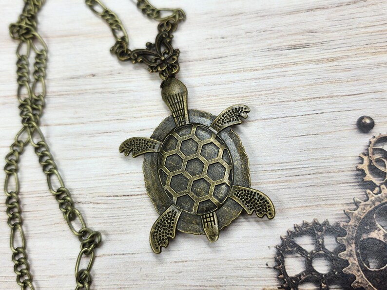 K&ouml;nnte beinhalten: Eine bronzefarbene Metallhalskette mit einem schildkr&ouml;tenf&ouml;rmigen Anh&auml;nger, der ein strukturiertes sechseckiges Panzermuster aufweist. Der Anh&auml;nger ist an einer dekorativen filigranen &Ouml;se befestigt und h&auml;ngt an einer passenden bronzefarbenen Kette.