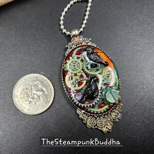 Peut inclure: Pendentif ovale avec un design steampunk repr&eacute;sentant deux corbeaux noirs, des engrenages et un fond orange et vert. Le pendentif est argent&eacute; avec des accents de roses. Le texte "TheSteampunkBuddha" est en bas.