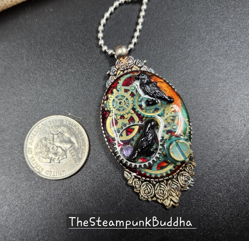 Peut inclure: Pendentif ovale avec un design steampunk repr&eacute;sentant deux corbeaux noirs, des engrenages et un fond orange et vert. Le pendentif est argent&eacute; avec des accents de roses. Le texte "TheSteampunkBuddha" est en bas.