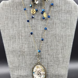 May include: A necklace and earring set with a steampunk theme. The necklace features an oval pendant with a fantasy image and a small unicorn charm. The earrings and necklace have blue crystal beads and bronze-colored metal accents.