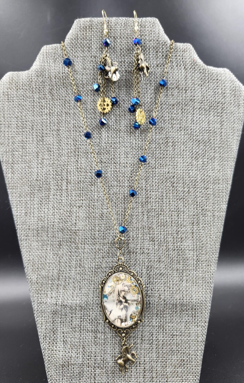 May include: A necklace and earring set with a steampunk theme. The necklace features an oval pendant with a fantasy image and a small unicorn charm. The earrings and necklace have blue crystal beads and bronze-colored metal accents.