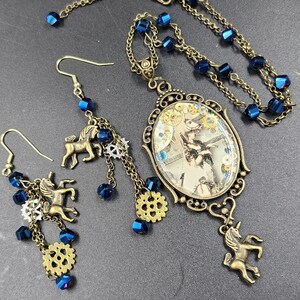 May include: A steampunk-style jewelry set featuring a necklace and earrings. The necklace has a pendant with a vintage image and a unicorn charm. The earrings have unicorn charms, blue beads, and gear accents. The set is made of bronze-toned metal.
