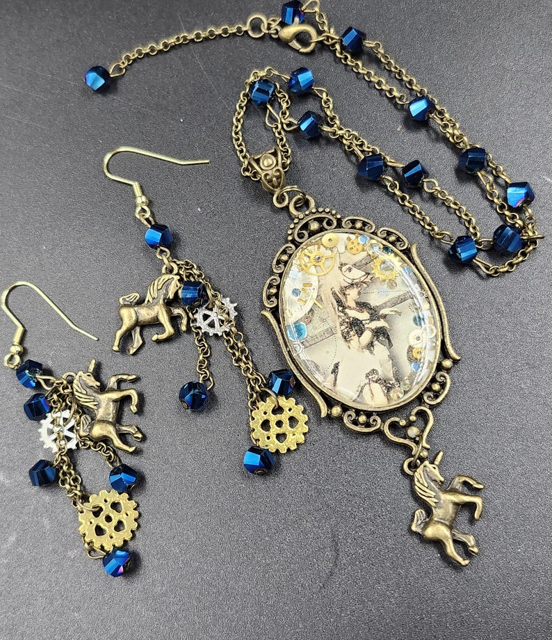 May include: A steampunk-style jewelry set featuring a necklace and earrings. The necklace has a pendant with a vintage image and a unicorn charm. The earrings have unicorn charms, blue beads, and gear accents. The set is made of bronze-toned metal.