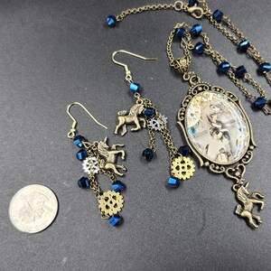 May include: A steampunk-style jewelry set featuring a necklace and earrings. The necklace has a pendant with a vintage image, and the earrings and pendant have bronze unicorn charms, blue beads, and gear accents. The set is on a dark gray surface.