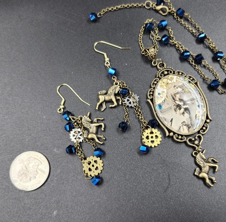 May include: A steampunk-style jewelry set featuring a necklace and earrings. The necklace has a pendant with a vintage image, and the earrings and pendant have bronze unicorn charms, blue beads, and gear accents. The set is on a dark gray surface.