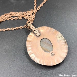 K&ouml;nnte beinhalten: Eine kupferfarbene Halskette mit Anh&auml;nger, geh&auml;mmert und mit einem ovalen Stein in der Mitte. Der Anh&auml;nger h&auml;ngt an einer feinen Kette. The Steampunk Buddha ist unten rechts aufgedruckt.