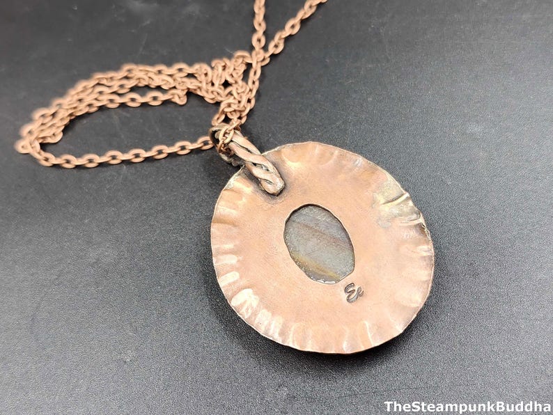 K&ouml;nnte beinhalten: Eine kupferfarbene Halskette mit Anh&auml;nger, geh&auml;mmert und mit einem ovalen Stein in der Mitte. Der Anh&auml;nger h&auml;ngt an einer feinen Kette. The Steampunk Buddha ist unten rechts aufgedruckt.
