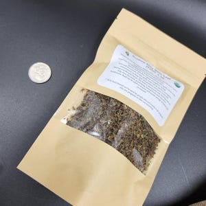 May include: A brown paper bag with a clear window containing dried herbs, next to a quarter. The bag has a white label with text that reads "Krishna Stay Holy Basil Leaf". The background is dark.