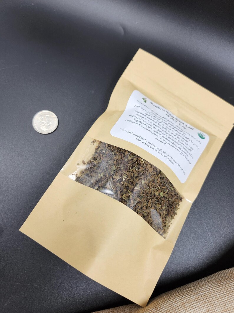 May include: A brown paper bag with a clear window containing dried herbs, next to a quarter. The bag has a white label with text that reads "Krishna Stay Holy Basil Leaf". The background is dark.