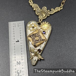 May include: A steampunk-style heart pendant necklace with a brass chain. The heart-shaped pendant features gears, a decorative square, and a dragonfly. The pendant is approximately 2 inches long. The text "TheSteampunkBuddha" is at the bottom.