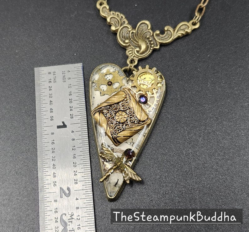 May include: A steampunk-style heart pendant necklace with a brass chain. The heart-shaped pendant features gears, a decorative square, and a dragonfly. The pendant is approximately 2 inches long. The text "TheSteampunkBuddha" is at the bottom.
