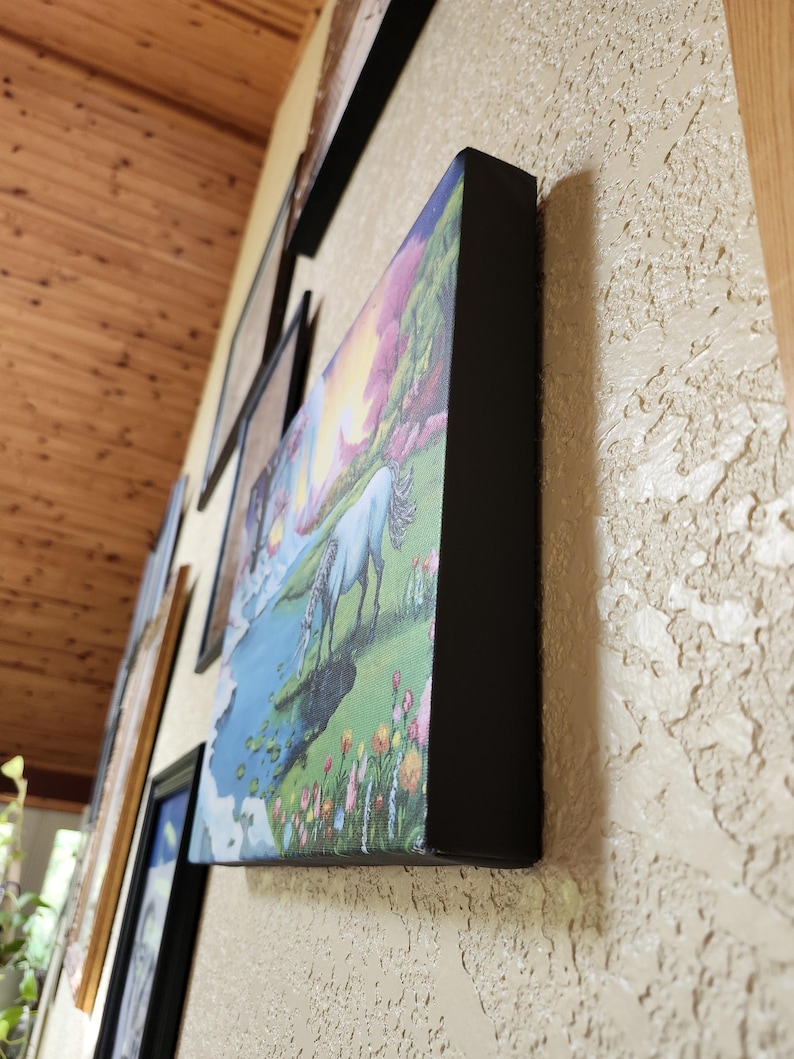 Puede incluir: Impresi&oacute;n en lienzo de un unicornio blanco bebiendo de un lago azul, rodeado de flores coloridas y un cielo de atardecer. La obra de arte est&aacute; enmarcada con un borde negro y montada en una pared beige texturizada.