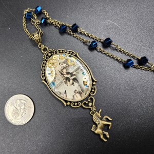 May include: An antique brass-toned necklace with a large oval pendant. The pendant features a vintage image with gears and blue accents. A small unicorn charm hangs below the pendant, and the necklace has blue bead accents.