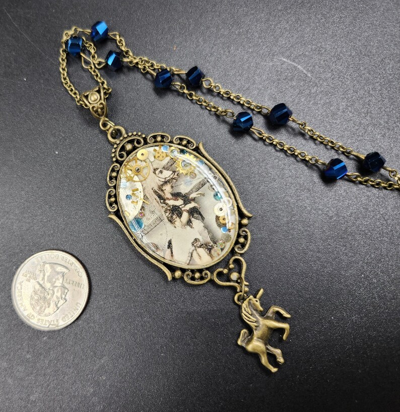 May include: An antique brass-toned necklace with a large oval pendant. The pendant features a vintage image with gears and blue accents. A small unicorn charm hangs below the pendant, and the necklace has blue bead accents.
