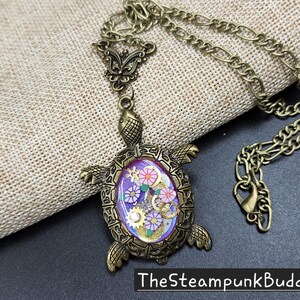 K&ouml;nnte beinhalten: Eine bronzefarbene Schildkr&ouml;ten-Halskette mit einem detaillierten Metallpanzer und Kopf. Die Mitte des Panzers enth&auml;lt eine violette Harzeinlage, die mit kleinen bunten Blumenformen und metallischen Zahnradkomponenten verziert ist. Der Text TheSteampunkBuddha ist sichtbar.