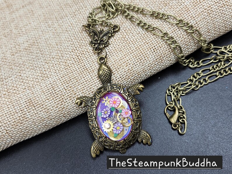 K&ouml;nnte beinhalten: Eine bronzefarbene Schildkr&ouml;ten-Halskette mit einem detaillierten Metallpanzer und Kopf. Die Mitte des Panzers enth&auml;lt eine violette Harzeinlage, die mit kleinen bunten Blumenformen und metallischen Zahnradkomponenten verziert ist. Der Text TheSteampunkBuddha ist sichtbar.