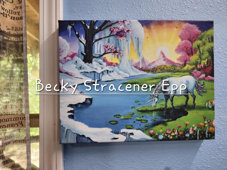 Puede incluir: Una obra de arte en lienzo enmarcada que representa un paisaje de fantas&iacute;a con un unicornio blanco bebiendo de un lago. La obra presenta una monta&ntilde;a nevada, &aacute;rboles rosas y el texto "Becky Stracener Epp".