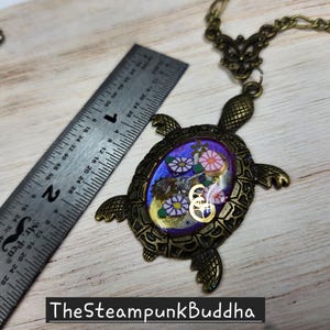 K&ouml;nnte beinhalten: Eine schildkr&ouml;tenf&ouml;rmige Anh&auml;ngerkette in Bronzefarbe mit einem violetten ovalen Zentrum, das florale Muster und metallische Zahnr&auml;der enth&auml;lt. Ein Metalllineal liegt neben dem Anh&auml;nger und zeigt, dass der Schildkr&ouml;tenk&ouml;rper etwa 5 cm lang ist.