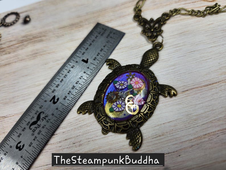 K&ouml;nnte beinhalten: Eine schildkr&ouml;tenf&ouml;rmige Anh&auml;ngerkette in Bronzefarbe mit einem violetten ovalen Zentrum, das florale Muster und metallische Zahnr&auml;der enth&auml;lt. Ein Metalllineal liegt neben dem Anh&auml;nger und zeigt, dass der Schildkr&ouml;tenk&ouml;rper etwa 5 cm lang ist.