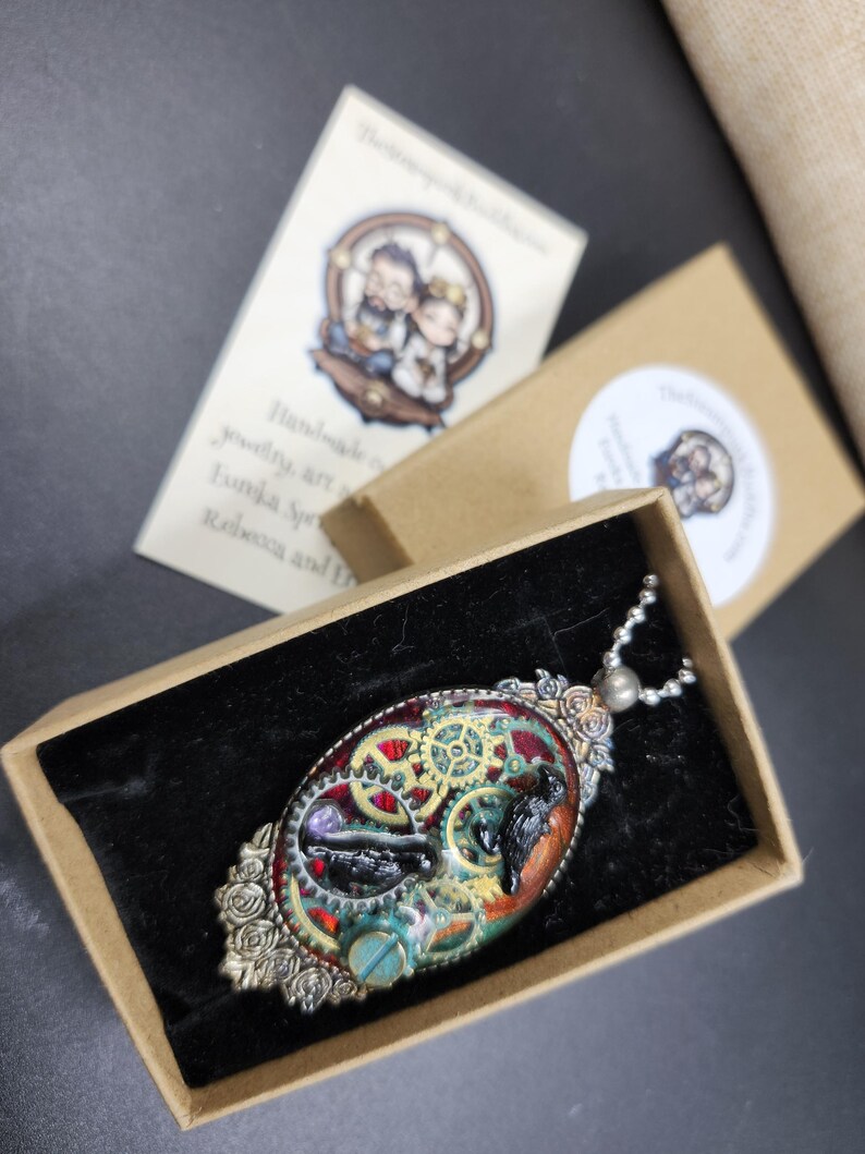 Peut inclure: Pendentif ovale avec un design steampunk, comprenant des engrenages, un oiseau et d'autres &eacute;l&eacute;ments dans une bo&icirc;te. Le pendentif a un cadre argent&eacute; avec des d&eacute;tails de roses et une cha&icirc;ne. La bo&icirc;te a un int&eacute;rieur en velours noir.