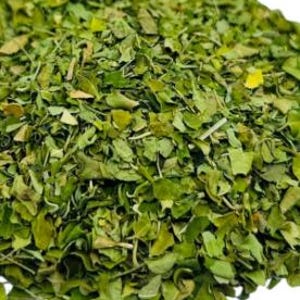 Op de afbeelding: Close-up van een hoop gedroogde groene moringabladeren. De bladeren zijn klein, met verschillende vormen en groentinten, en lijken fijngehakt. De afbeelding is goed belicht en toont de textuur en details van de bladeren.