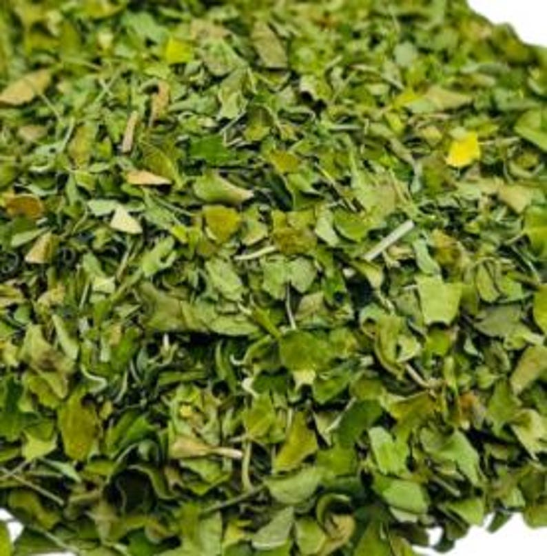 Op de afbeelding: Close-up van een hoop gedroogde groene moringabladeren. De bladeren zijn klein, met verschillende vormen en groentinten, en lijken fijngehakt. De afbeelding is goed belicht en toont de textuur en details van de bladeren.