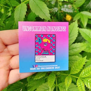 May include: A pink enamel pin featuring a cartoon character on a blue background. The character is a pink animal with a yellow head and blue eyes. The pin reads "Fuchsia 09-19 Precious" and "Uncommon Nonsense" with a social media handle. The pin has a gradient background with pink at the top and blue at the bottom.